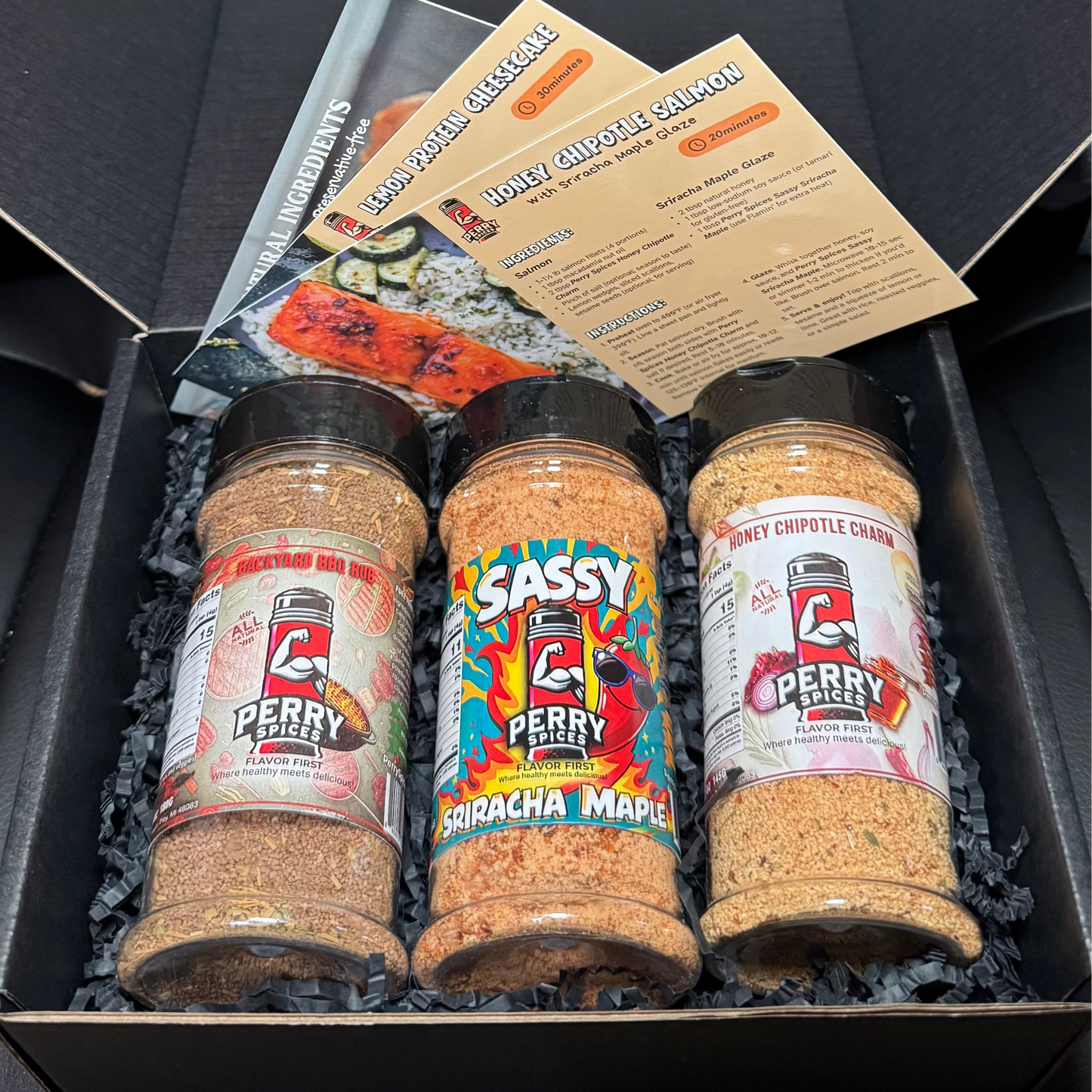 Three jars of flavorful seasonings in a gift box with a 3 recipe cards on a dark surface