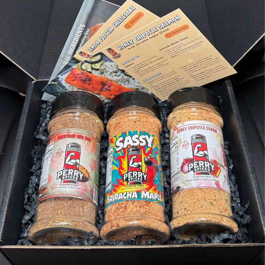 Three jars of flavorful seasonings in a gift box with a 3 recipe cards on a dark surface
